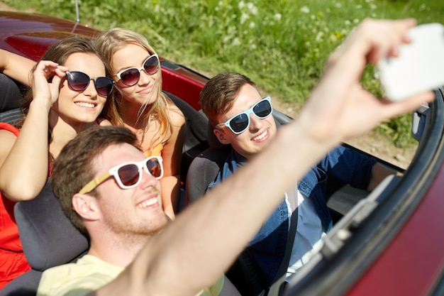 Freizeit-, Roadtrip-, Reise- und People-Konzept - glückliche Freunde, die im Cabriolet fahren und Selfie per Smartphone im Freien machen