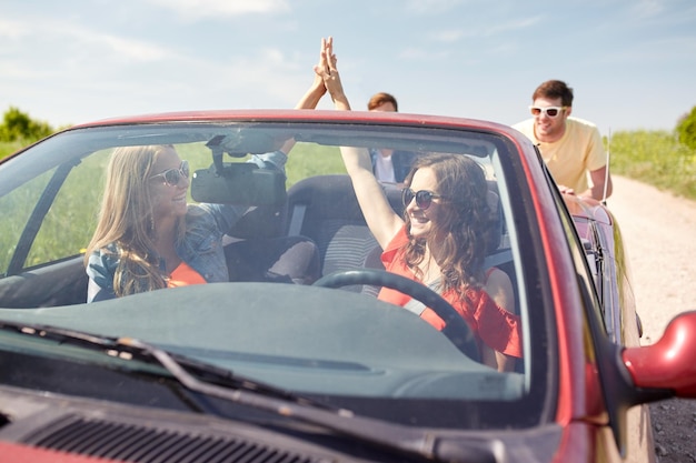 Freizeit-, Straßenreise-, Reise- und Personenkonzept - glückliche Freunde, die kaputtes Cabriolet-Auto entlang der Landstraße schieben