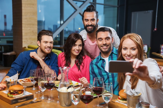 freizeit-, technologie-, freundschafts-, menschen- und urlaubskonzept - glückliche freunde, die zu abend essen und selfie per smartphone im restaurant machen