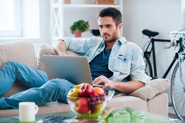 Freizeit zu Hause genießen. Schöner junger Mann, der am Laptop arbeitet, während er zu Hause auf der Couch liegt