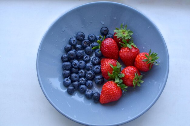 Foto fresas y arándanos en un plato azul en el alféizar de la ventana