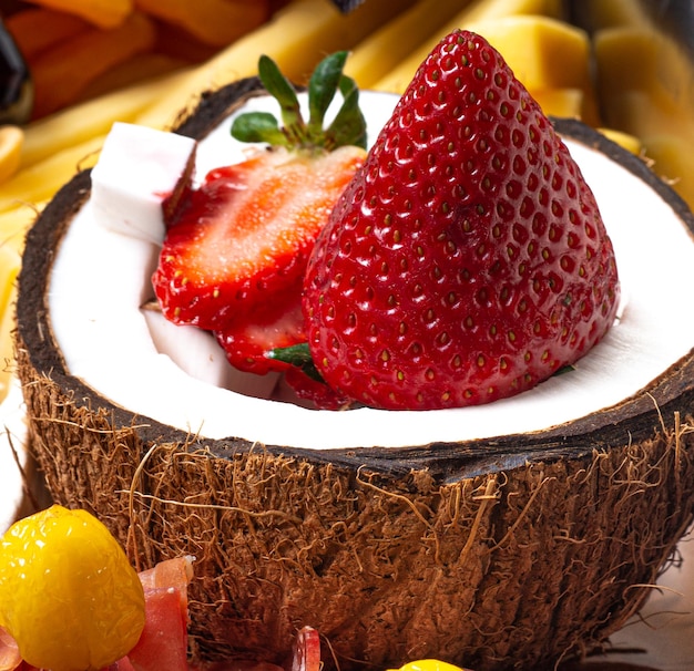 Fresas dentro de un coco en rodajas sobre una mesa de mármol decorada con frutas y embutidos alrededor.