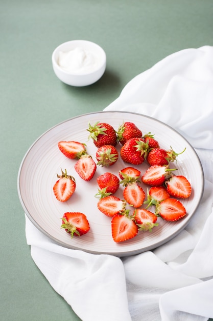 Fresas frescas enteras y en rodajas en un plato y crema agria en un recipiente sobre una mesa verde.