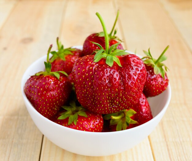 Fresas frescas en un recipiente blanco sobre una mesa de madera clara. De cerca