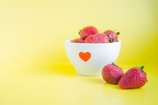 Fresas frescas en un tazón blanco con corazón rojo sobre el fondo amarillo brillante con copyspace