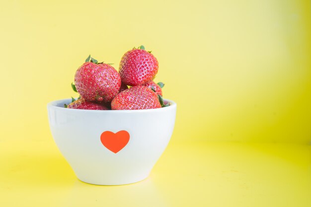 Fresas frescas en un tazón blanco con corazón rojo sobre el fondo amarillo brillante con copyspace