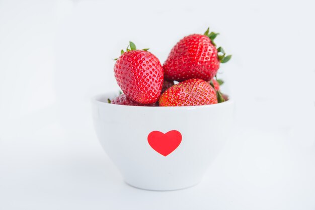 Fresas frescas en un tazón blanco con corazón rojo sobre el fondo blanco de cerca