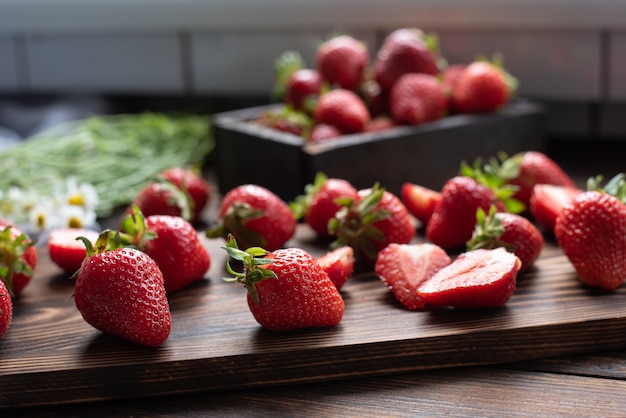 Fresas maduras sobre una tabla de cortar de madera en la cocina con un ramo de margaritas, concepto de verano, de cerca.