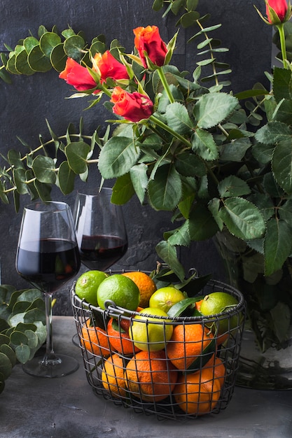 Fresas y moras en un plato verde y mandarinas en una cesta cerca de un jarrón con un ramo de rosas, sobre un fondo negro. dos copas de vino tinto