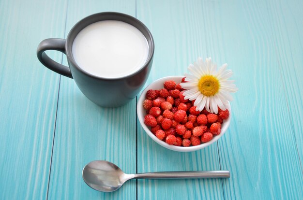 Foto fresas silvestres en tazón de fuente blanco, leche en taza y cuchara de plata sobre fondo de madera azul, vista superior. espacio para texto. delicioso postre de verano elaborado con aromáticos frutos del bosque. desayuno saludable