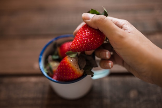 Fresas en una taza de esmalte blanco y una mano sosteniendo una fresa