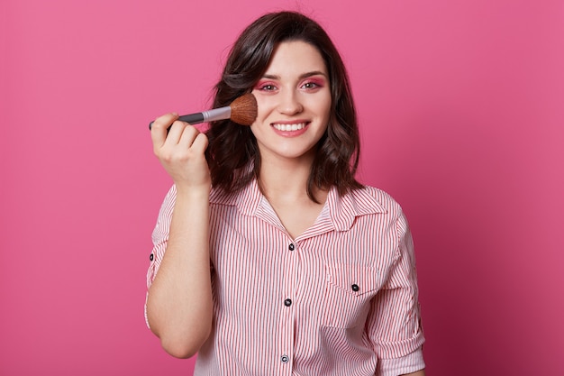 Fresca, encantadora y encantadora joven modelo femenina sostiene un cepillo cosmético, sonríe ampliamente, tiene una piel sana