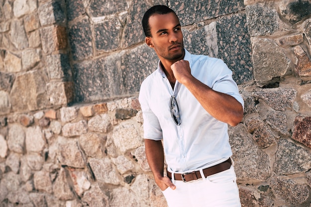 Fresco y elegante. Apuesto joven africano en ropa casual elegante ajustando el cuello de su camisa mientras está de pie contra la pared de piedra al aire libre