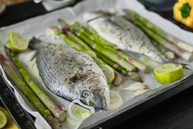 Fresco eviscerado y sazonado con sal y sal listo para hornear dorado en una bandeja para hornear cubierta con papel de hornear con espárragos, lima, ajos tiernos y cebollas.