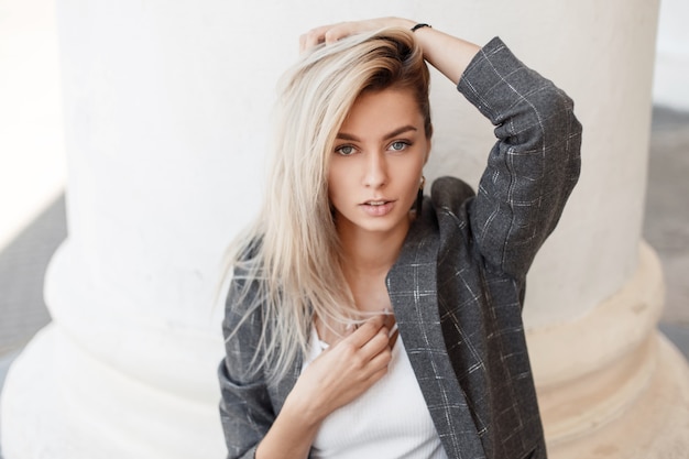 Foto fresco retrato de una bella mujer joven con una chaqueta gris de moda con una camiseta blanca sentada cerca de una columna blanca en la calle y mirar a la cámara