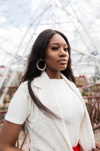 Fresco retrato joven mujer negra con ojos increíbles con labios sexy con piel limpia y saludable en ropa blanca elegante en el cielo y la noria en el parque. Hermosa chica africana se encuentra cerca de paseos en la calle.