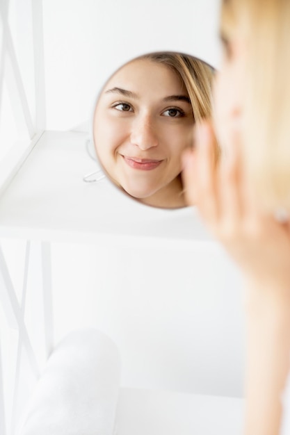 Frescura de belleza Dermatología femenina Salud de la piel Mujer alegre tocando la hermosa cara radiante en el reflejo del espejo en el espacio libre de luz