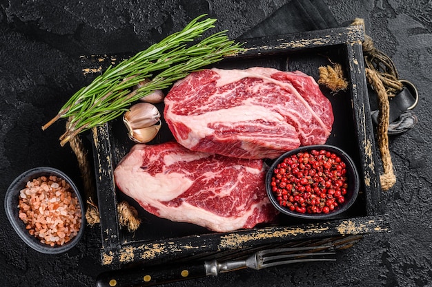 Fresh Raw Chuck Roll Zarte Rindersteaks in einem Holztablett mit Kräutern Schwarzer Hintergrund Draufsicht