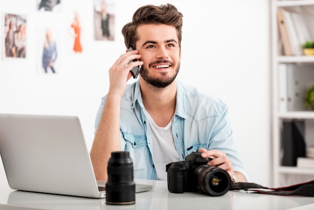 Freude am kreativen Arbeiten. Glücklicher junger Mann, der am Handy spricht und lächelt, während er an seinem Arbeitsplatz sitzt und eine Digitalkamera hält holding