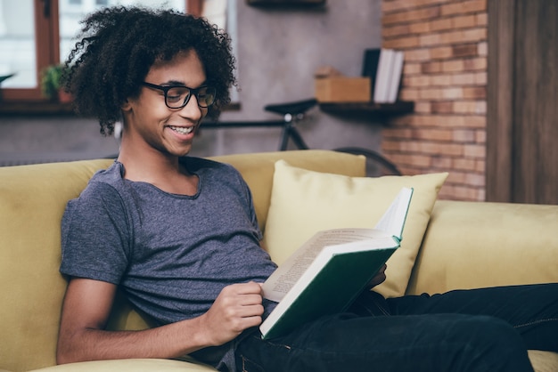 Freude am neuen Kapitel. Seitenansicht eines fröhlichen jungen afrikanischen Mannes, der ein Buch mit Lächeln liest und eine Brille trägt, während er zu Hause auf der Couch sitzt