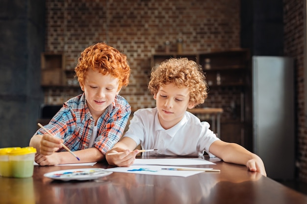 Freudig konzentrierte Kinder sitzen nebeneinander und konzentrieren ihre Aufmerksamkeit auf Papier, während sie kreativ werden und gemeinsam etwas Interessantes malen.