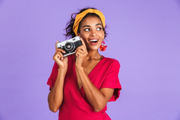 Freudige afrikanische Frau im Kleid, die Retro-Kamera hält und über lila Wand wegschaut