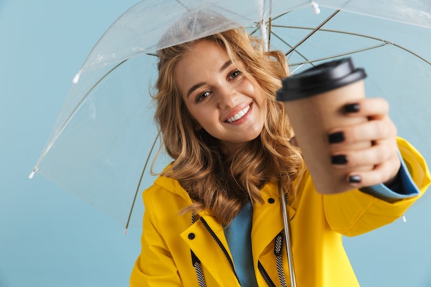 freudige Frau 20s tragen gelben Regenmantel, der unter transparentem Regenschirm mit Kaffee zum Mitnehmen steht