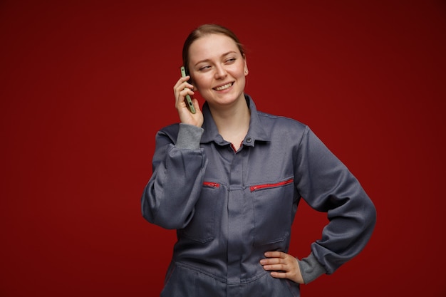 Freudige junge blonde Ingenieurin in Uniform, die am Telefon spricht und die Hand auf der Taille hält und die Seite lächelnd betrachtet