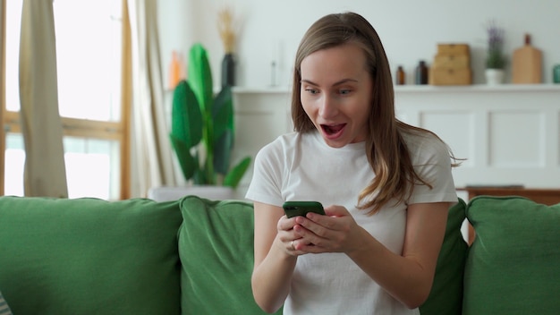 Freudige junge Dame, die auf der Couch sitzt, mit Smartphone.