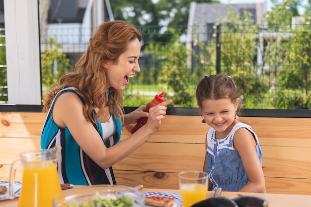 Freudige junge Frau, die eine Flasche Ketchup hält, während sie mit ihrer Tochter spielt