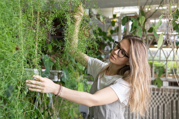 Freudige junge Gärtnerin im Pfandrechtkleid, das üppige Spargelfarn-Zimmerpflanze berührt. Grün zu Hause. Liebe zu Pflanzen. Gemütlicher Innengarten.