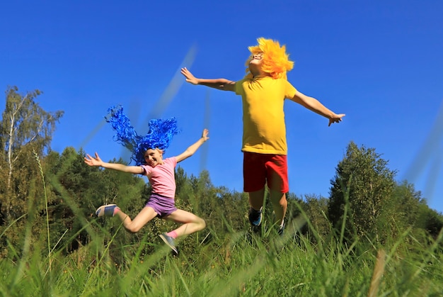 Freudige Kinderjungen und -mädchen mit blauen und gelben Haaren, die auf Rasen in der Natur nahe dem Wald bei sonnigem Wetter springen.