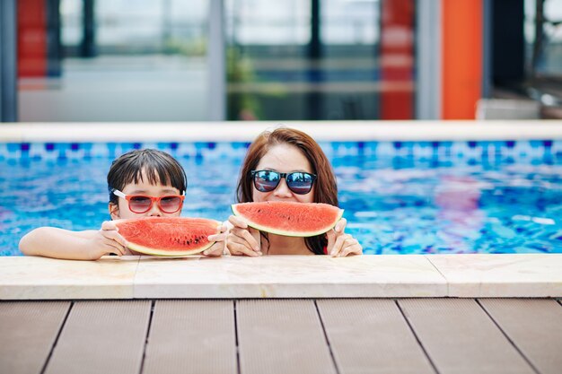 Freudige Mutter und kleine Tochter in der Sonnenbrille, die im Schwimmbad steht und frische Wassermelone isst