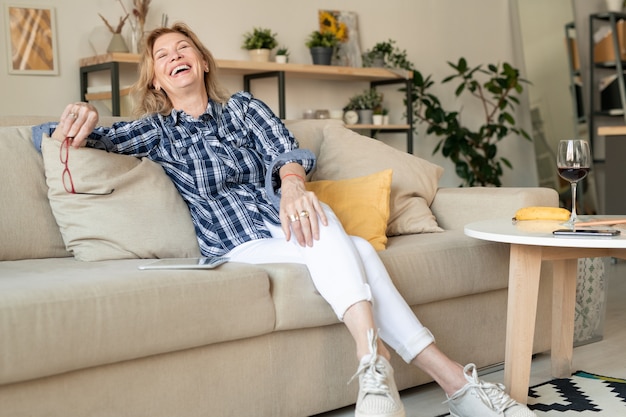 Freudige reife Frau in der Freizeitkleidung, die lacht, während sie Tag zu Hause verbringt und auf der Couch am kleinen Tisch mit Glas Rotwein entspannt