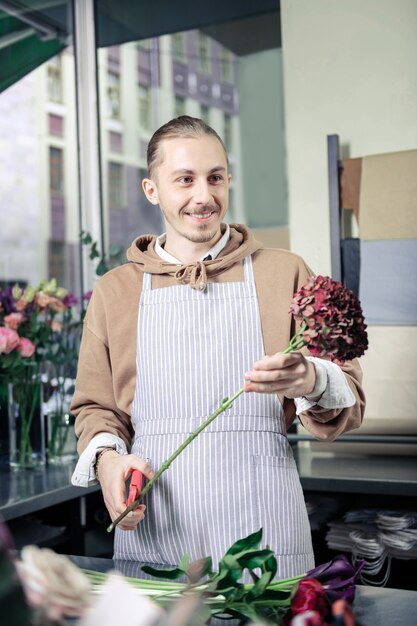 Freudige Stimmung. Freundlicher junger Florist, der Positivität ausdrückt, während Blumen für Blumenstrauß vorbereitet