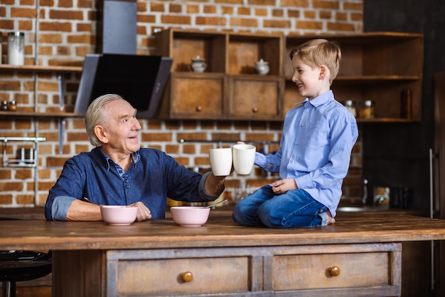 Freudiger älterer Mann, der Tee trinkt, während er mit seinem Enkel frühstückt