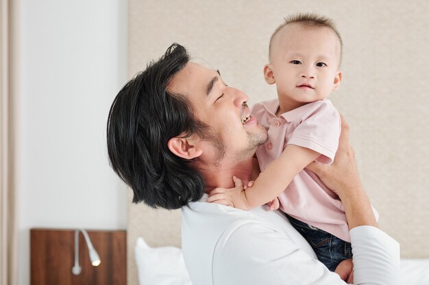 Freudiger junger asiatischer Mann, der seinen kleinen neugeborenen Sohn trägt und ihn mit Liebe und Zärtlichkeit ansieht