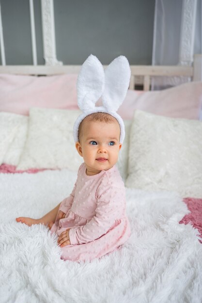 Freudiges Baby in einem rosa Kleid und einem Stirnband mit Hasenohren sitzt auf dem Bett im Zimmer