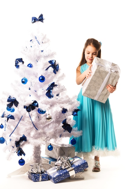 Freudiges junges Mädchen in einem blauen Kleid und hält ein Geschenk in ihren Händen und schmückt einen weißen künstlichen Weihnachtsbaum des neuen Jahres auf einer weißen Wand