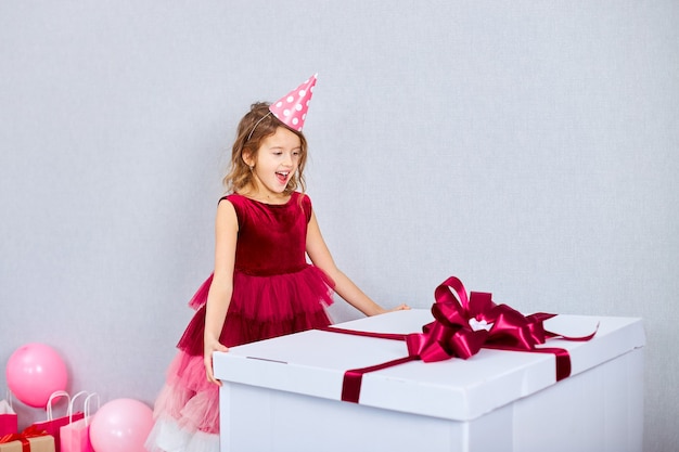 Freudiges kleines Mädchen im rosa Kleid und im Hut öffnet große Geschenkbox mit Luftballons