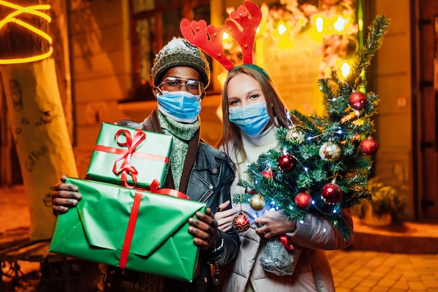 Freudiges paar, das am abend in der stadt mit weihnachtsgeschenken in medizinischen masken steht