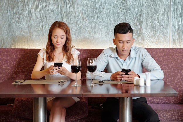 Freund und Freundin trinken Wein, warten auf Essen und checken Social Media auf Smartphones