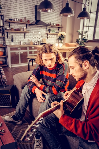 Freund zuhören. Blonder Mann mit gestreiftem Pullover, der seinem Freund beim Gitarrespielen zuhört playing