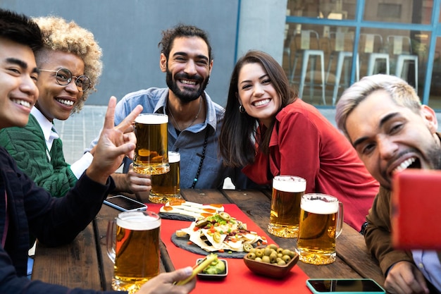 Foto freunde aus verschiedenen rassen machen ein selfie mit dem handy, während sie kühles bier und essen in einer bar im freien genießen