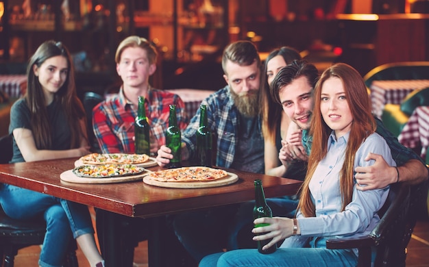 Freunde, die in einer Bar etwas trinken, sitzen an einem Holztisch mit Bieren und Pizza.