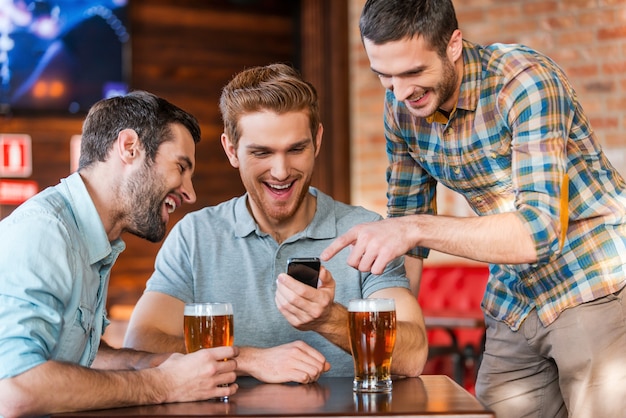 Freunde, die Spaß haben. Drei glückliche junge Männer in Freizeitkleidung trinken Bier in der Kneipe, während einer von ihnen auf das Smartphone zeigt und lächelt