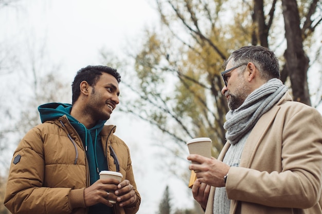 Freunde, ein Senior und ein junger Mann, die im HerbstparkxA zusammen spazieren gehen und reden und Kaffee trinken