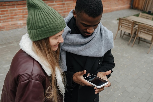 Freunde hängen auf der Straße herum und schauen auf einen Telefonbildschirm. Beide kleideten sich warm für den Spätherbst. Interracial Freundschaftskonzept des gegensätzlichen Geschlechts.