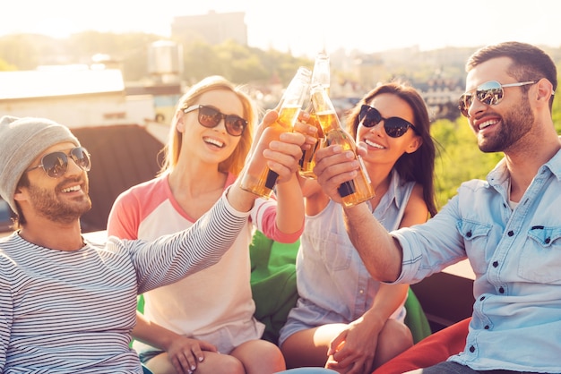 Freunde und Bier. Vier junge fröhliche Leute, die mit Bier jubeln und lächeln, während sie an den Sitzsäcken auf dem Dach des Gebäudes sitzen