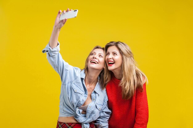 Freundinnen, die Zeit genießen Porträt von zwei schönen Frauen in stilvoller Kleidung, die mit einem zahnigen Lächeln stehen und Selfie am Telefon machen, glückliche Erinnerungen, Studioaufnahme isoliert auf gelbem Hintergrund